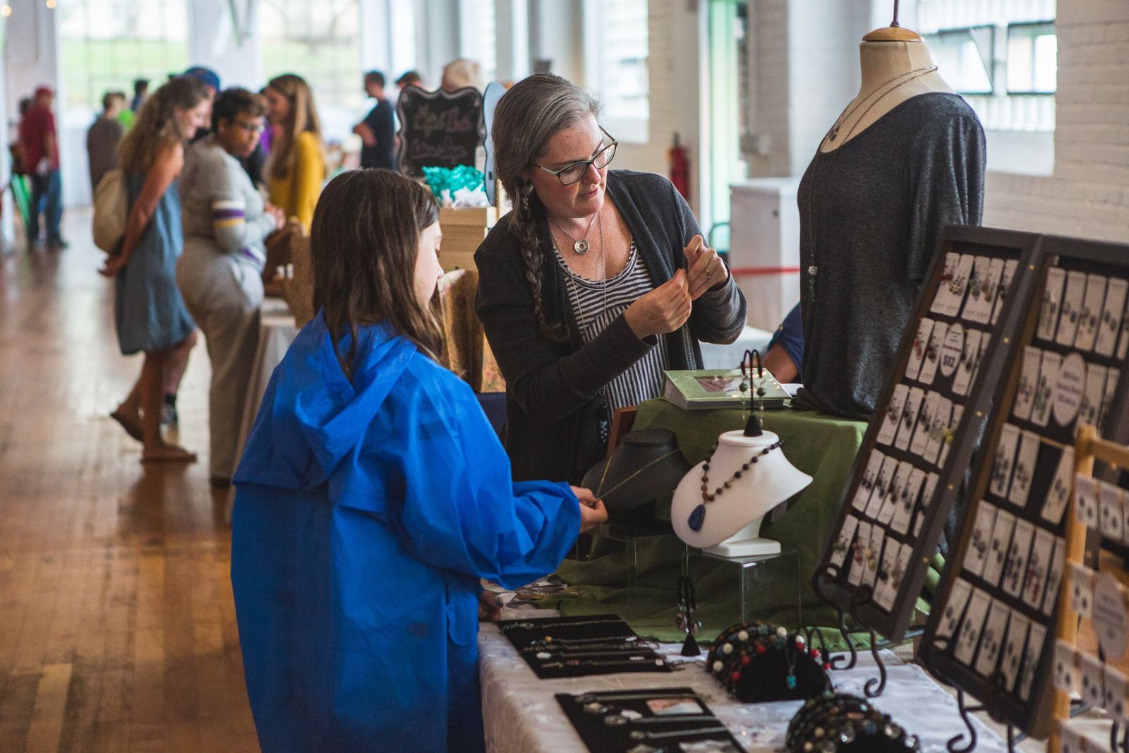A vendor with glasses and a braided ponytail shows a necklace to a girl in a blue raincoat at a jewelry display, with shoppers browsing booths along a wood-floored hall behind them.