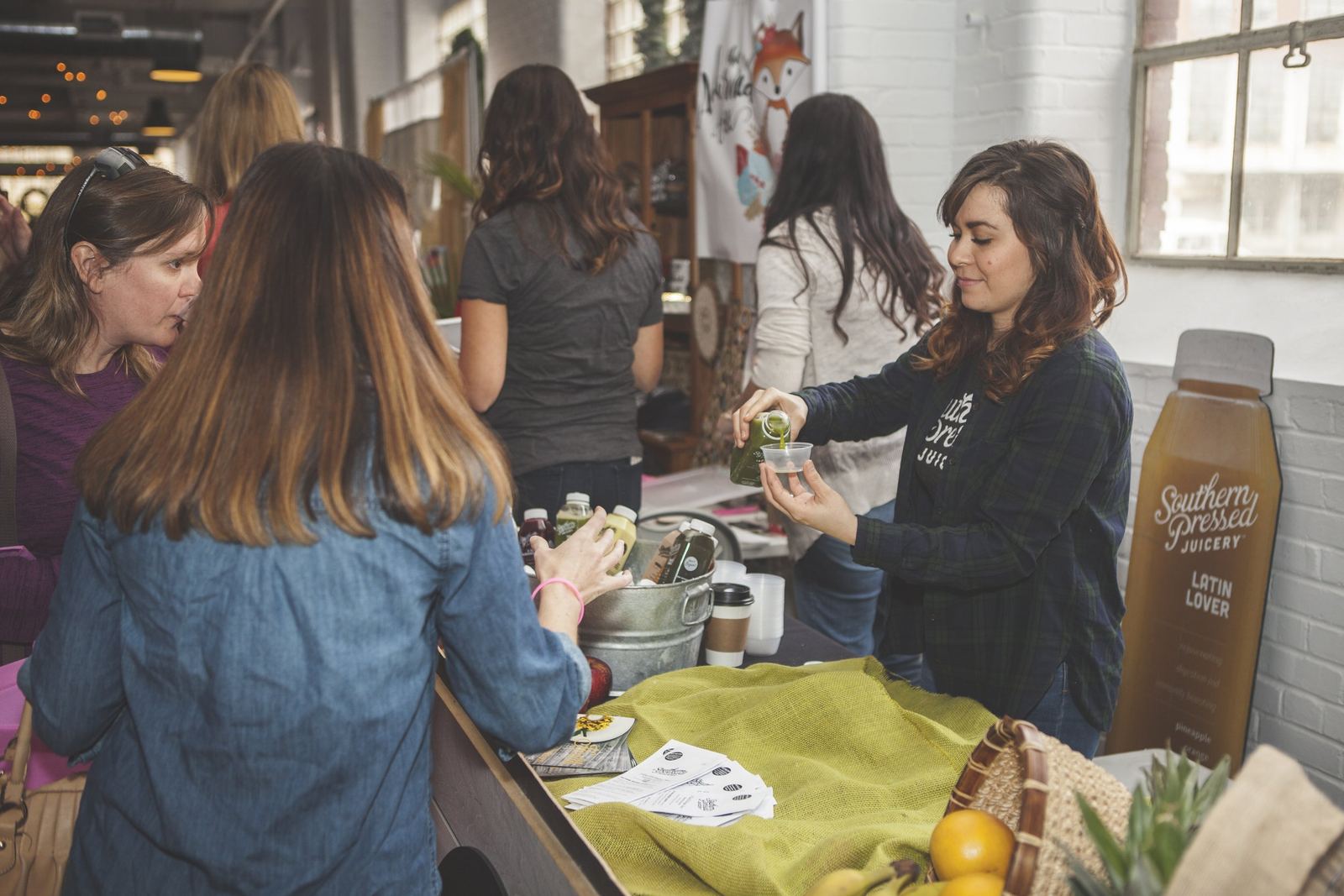 A woman at a juice stand pours a green juice into a small cup for a customer while others browse the selection of juices and smoothies.