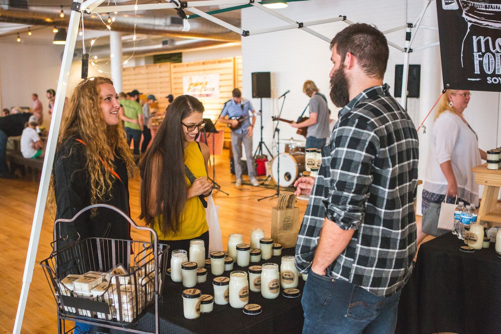 People at a market stall with jars of cream or yogurt, engaging in conversation, with an indoor event space in the background including musical performers.